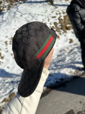 Svart Gucci keps med GG-mönster - Säljer en svart keps från Gucci med klassiskt GG-monogram över hela kepsen och den ikoniska gröna och röda randen på sidan. Kepsen har böjd skärm och justerbar rem bak. Perfekt för dig som vill ha en stilren och lyxig accessoar. Priset går att diskutera vid snabb affär och lappen ligger bredvid tog bort den. Väldigt bra skick inga skador osv