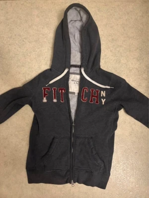 Mörkgrå hoodie Abercrombie & Fitch - Archive Vintage Mörkgrå hoodie med dragkedja från Abercrombie & Fitch. Stor broderad text 'FITCH NY' i rött och vitt på bröstet, vita snören i huvan och två fickor framtill. Mjuk insida och klassisk passform. Använd med i mycket bra skick. 