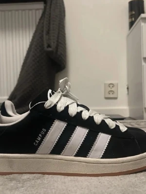 Adidas Campus svartvita sneakers - Säljer ett par klassiska Adidas Campus sneakers i svart mocka med vita läderdetaljer och de ikoniska tre ränderna på sidan. Skorna har vita skosnören och en låg siluett med gummisula. Perfekta för en clean och tidlös streetstil.