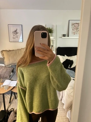 Grön oversized stickad tröja - Säljer en grön oversized stickad tröja med ljusrosa detaljer vid hals, ärmslut och nederkant. Tröjan har en lös passform och bred urringning som kan bäras off-shoulder. Suuuper snygg nu till våren. Använd fåtal gånger!💚🩷