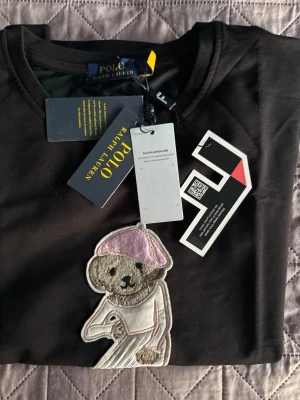Svart t-shirt Polo Ralph Lauren björn - Svart t-shirt från Polo Ralph Lauren med en stor broderad björn på bröstet, klädd i ljusrosa och vita kläder. Klassisk rund hals och korta ärmar. Perfekt för dig som gillar ikoniska detaljer och vill sticka ut lite extra.