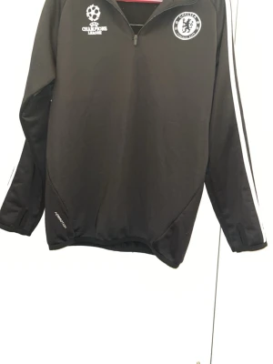 Chelsea FC svart träningsjacka - Snygg svart träningsjacka från Adidas med Chelsea FC-logga och Champions League-märke på bröstet. Jackan har halv dragkedja, vita ränder längs ärmarna och är tillverkad i ett sportigt funktionsmaterial. Perfekt för fotbollsträning eller chill.