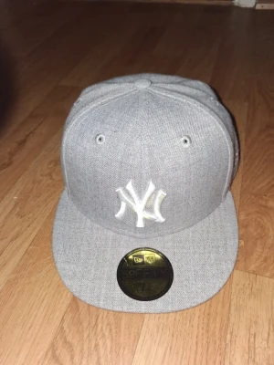 Grå New Era Yankees keps - Snygg grå snapback-keps från New Era med New York Yankees-logga broderad framtill i vitt. Klassisk flat brim och MLB-logga baktill. Tillverkad i ett mjukt tygmaterial som ger en clean och stilren look. Perfekt för dig som gillar streetwear och sportig stil.