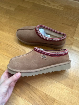 Bruna UGG regenerate tasman - Säljer ett par bruna UGG tofflor i mocka med dekorativ röd och vit kant runt öppningen. Tofflorna har en rejäl beige sula och UGG-logga på sidan och hälen. Perfekta för att hålla fötterna varma och snygga hemma.