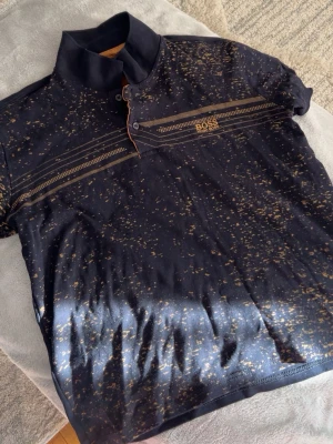 Skjorta  - Säljer en svart Henley-skjorta från BOSS med unikt guldmönster och knappar vid halsen. Skjortan har korta ärmar och detaljerade linjer över bröstet samt BOSS-logga i guld. Perfekt för dig som vill sticka ut med en cool och modern look. Kan diskutera pris. Storlek M-L men står XL, däremot liten.