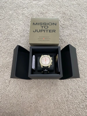 Omega x Swatch Mission to Jupiter - Snygg Omega x Swatch Speedmaster MoonSwatch Mission to Jupiter med beige boett och ljus urtavla. Klockan har svarta detaljer och en svart textilrem med Omega-logga. Perfekt för dig som gillar unik design och ikoniska samarbeten.