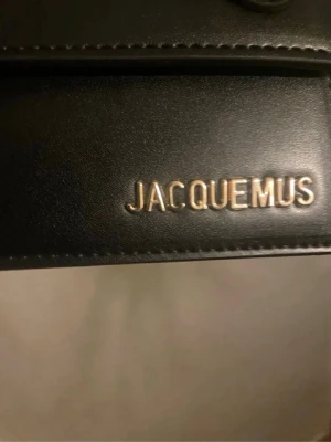 Jacquemus axelväska/handväska  - Snygg svart axelväska från Jacquemus i slätt läder med guldfärgad logga framtill. Väskan har ett stilrent och minimalistiskt uttryck med ett rejält handtag och raka linjer. Perfekt för dig som gillar trendiga accessoarer och vill ha något som sticker ut.