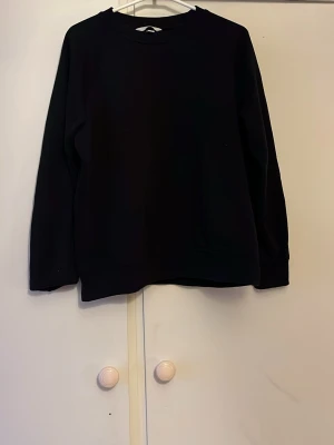 Svart basic hoodie utan tryck - En enkel svart hoodie i mjukt material, perfekt för en clean och avslappnad look. Modellen har rund halsringning och långa ärmar. Inga synliga loggor eller detaljer, vilket gör den lätt att matcha med allt.