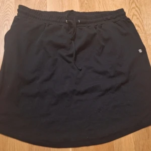 Svart kortkjol med snörning - Svart kortkjol med snörning i midjan och en liten logotyp på sidan. Kjolen har en enkel och avslappnad design, perfekt för att matcha med olika toppar. Materialet ser ut att vara mjukt och bekvämt, troligen bomull. Passformen är normal och kjolen slutar ovanför knäna.