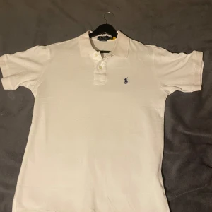 Vit piké från Polo Ralph Lauren - Klassisk vit pikétröja från Polo Ralph Lauren med blå broderad logga på bröstet. Tröjan har korta ärmar, krage och knappar framtill. Perfekt basic-plagg med snygg passform och tidlös stil.