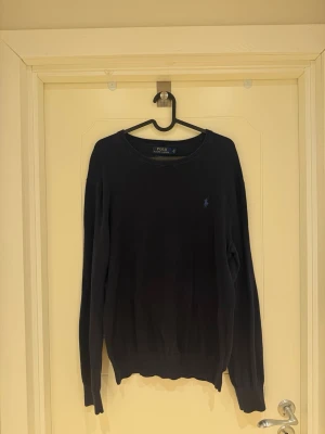 Mörkblå tröja Polo Ralph Lauren - Snygg mörkblå stickad tröja från Polo Ralph Lauren med rund halsringning och diskret broderad logga på bröstet. Tröjan har långa ärmar och ribbade muddar vid ärmslut och nederkant. Perfekt för lager på lager och stilrena outfits. Materialet är Pima Cotton.        Modellen är 185cm lång och väger 82kg.
