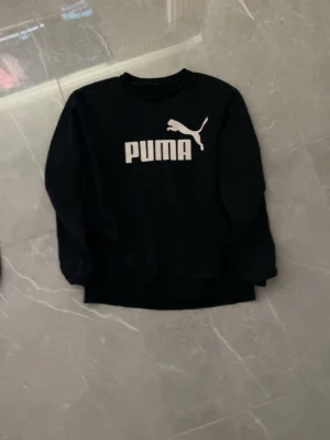 Svart sweatshirt från Puma - Säljer en svart sweatshirt från Puma med stort vitt logotryck på bröstet. Tröjan har rund halsringning och långa ärmar. Klassisk och enkel design som passar till det mesta. Materialet känns mjukt och bekvämt mot huden.