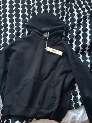 Svart Essentials hoodie Fear of God - Svart hoodie från Fear of God Essentials med broderad logga på bröstet och ryggen. Klassisk huva, stor frontficka och ribbade muddar. Materialet är mjukt och känns riktigt skönt mot huden. Perfekt för dig som gillar streetwear och minimalistisk stil.