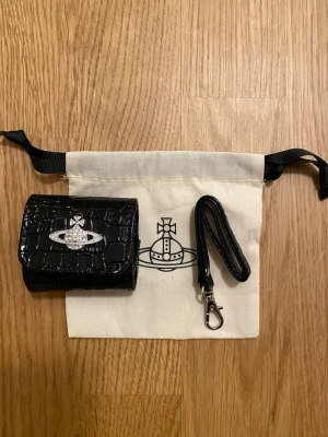 Svart Vivienne Westwood airpods case - Snygg svart airpods case i croco-mönstrat konstläder från Vivienne Westwood. Har ett silverfärgat emblem framtill och medföljande handledsrem med karbinhake. Kommer med original tygpåse med logga.