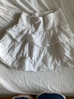 Vit volangkjol från Gina Tricot - Supersöt vit kortkjol från Gina Tricot med volanger och smock i midjan. Kjolen har broderade detaljer och är gjord i ett lätt bomullstyg som passar perfekt till varma dagar. Enkel att matcha med både toppar och sneakers för en fräsch look.