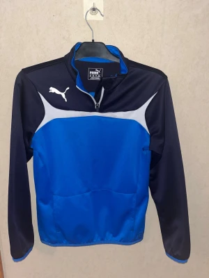 Blå och svart träningsjacka Puma - Sportig träningsjacka från Puma i blått och svart med vita detaljer. Jackan har halv dragkedja, lång ärm och klassisk Puma-logga på bröstet. Tillverkad i ett lätt och funktionellt material som passar perfekt till träning eller uppvärmning.