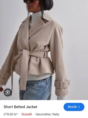 Beige kort kappa med bälte från Nelly - Beige kort kappa från Nelly med bälte i midjan och dekorativa spännen vid ärmsluten. Perfekt nu till våren🌷Den är i storlek S/M men enligt mig är den liten i storleken. Jag säljer den för 170kr, men är öppen för prisförslag<3