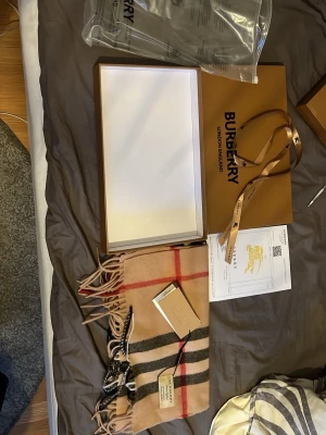 Beige klassisk halsduk från Burberry - Säljer en ikonisk beige halsduk från Burberry i mjuk ull med det klassiska rutmönstret i svart, vitt och rött. Halsduken har fransar i ändarna och kommer med originalförpackning och taggar kvar. Perfekt accessoar för att lyfta din outfit.