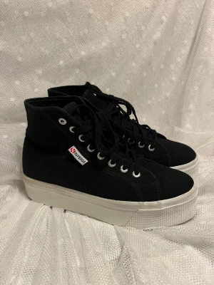 Svarta Superga platåsneakers - Svarta sneakers från Superga med hög platåsula i vitt. Skorna har canvasöverdel, svarta skosnören och metallfärgade öljetter. Klassisk Superga-logga på sidan och en clean, tidlös design som passar till det mesta. Stl 40. Använda två gånger. Rök och Djurfritt.