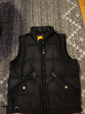 Svart dunväst från Parajumpers - Snygg svart dunväst från Parajumpers med hög krage och gul detalj vid halsen. Västen har två stora framfickor med dragkedja och tryckknapp samt robust dragkedja framtill. Perfekt för lager på lager och streetwear-vibe.