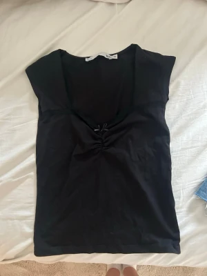 Svart topp med rosett från 157 - Snygg svart topp från 157 med liten rosett och rynkdetalj vid bysten. Toppen har korta ärmar och en djupare v-ringning med spetskant. 