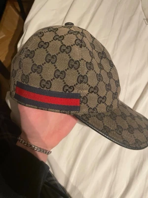 Gucci beige keps med GG-mönster - Säljer en beige keps från Gucci med det klassiska GG-monogrammet över hela kepsen. På sidan finns en ikonisk röd och marinblå rand. Kepsen har böjd skärm och justerbar rem bak. Materialet är canvas med detaljer i läder.