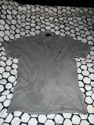 Grå pikétröja från Polo Ralph Lauren - Klassisk grå pikétröja från Polo Ralph Lauren med korta ärmar och liten mörkblå logga broderad på bröstet. Tröjan har krage och knappar vid halsen. Perfekt till en avslappnad och stilren look.