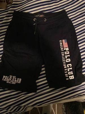 Svarta shorts från Polo Club - Snygga svarta shorts från Polo Club Vincent Collection med tryckt logga och amerikansk flagga på ena benet. Shortsen har snörning i midjan och sidofickor, perfekta för en avslappnad stil. Tillverkade i mjukt material för extra komfort.