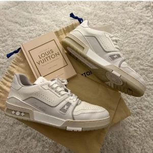 Louis Vuitton vita sneakers - Stilrena vita Louis Vuitton sneakers med grå detaljer och perforerade partier. Skorna har en chunky sula, klassisk snörning och diskreta loggor på sidan och sulan. Tillverkade i läder och mesh för en exklusiv känsla och modern look.