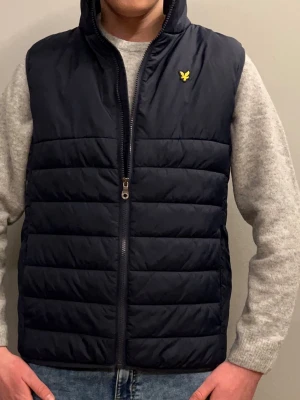 Mörkblå dunväst från Lyle & Scott - Snygg marinblå dunväst från Lyle & Scott med gul logga på bröstet. Västen har hög krage, dragkedja framtill och quiltad design. Perfekt att slänga över en hoodie eller tröja för extra värme. Klassisk och stilren look som funkar till det mesta. Nyskick, sitter som S-XS i storlek. Hör gärna av dig vid frågor eller tankar. 🤗