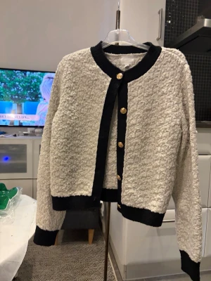 Beige bouclékofta med svarta detaljer - Säljer en beige bouclékofta med svarta kanter och guldknappar framtill. Koftan har rund halsringning, långa ärmar och en croppad passform. Materialet är strukturerat och ger en lyxig känsla. Perfekt för dig som gillar klassisk men trendig stil.