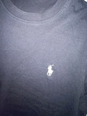 Svart t-shirt från Ralph Lauren - Klassisk svart t-shirt från Ralph Lauren med den ikoniska vita polospelaren broderad på bröstet. T-shirten har rund halsringning och korta ärmar. Tillverkad i mjuk bomull för en skön och avslappnad stil.