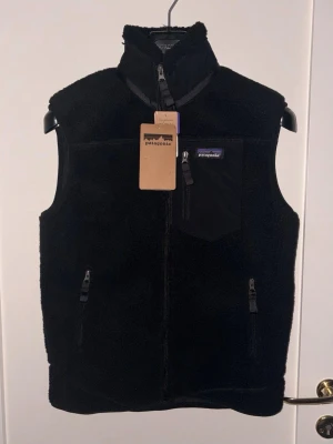 Svart fleeceväst från Patagonia - Säljer en svart fleeceväst från Patagonia med hög krage, dragkedja framtill och tre fickor med dragkedja. Västen har en bröstficka i nylon med Patagonias logga och är gjord i mjukt, värmande fleecematerial. Perfekt för lager-på-lager och outdoor.