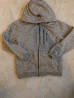 Grå hoodie från Sail Racing - Snygg ljusgrå hoodie från Sail Racing med dragkedja framtill och en praktisk bröstficka med zip. Hoodien har en broderad logga på huvan och tryck på bröstet. Tillverkad i mjukt bomullsmaterial och har ribbade muddar vid ärmar och nederkant.