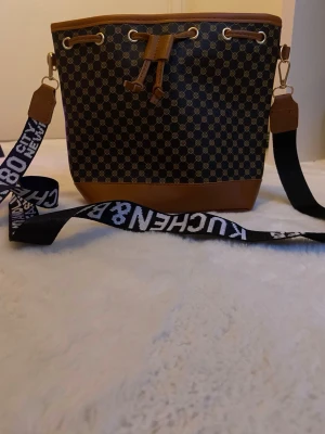 Brun bucket bag med svart axelband - Cool brun handväska i bucket-modell med svart innerfoder och bredt axelband med vit text. Väskan har metallringar och snörning upptill, perfekt för att bära dina essentials. Materialet är troligtvis konstläder och textil.