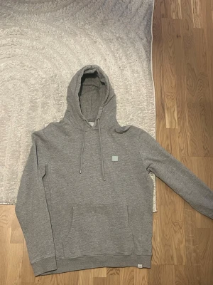 Grå hoodie från Les deux - Säljer en stilren grå hoodie från Les deux med broderad logga på bröstet. Hoodien har stor huva med dragsko och en klassisk känguruficka framtill. Tillverkad i mjukt bomullsmaterial som är skönt mot huden. Perfekt för en avslappnad och trendig look.
