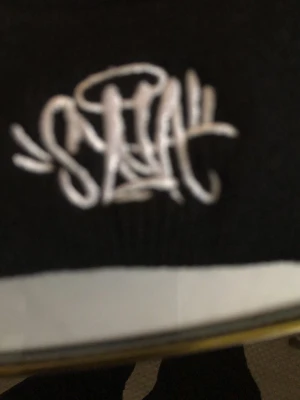 Svart mössa med STAY brodyr - Säljer en svart mössa med uppvikt kant och vit broderad STAY-logga framtill i graffiti-stil. Mössan är ribbstickad och passar perfekt för dig som vill ha en clean streetwear-look. Materialet känns mjukt och stretchigt, vilket gör den bekväm att bära.