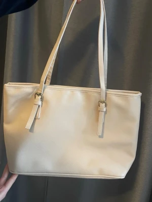 Beige shoppingväska med långa handtag - Stilren shoppingväska i beige konstläder med långa axelband och guldfärgade spännen. Väskan har en rymlig insida med dragkedjefack och tryckknapp för stängning. Perfekt för dig som vill ha plats för allt du behöver under dagen.