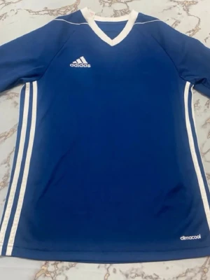 Blå Adidas träningströja Climalite - Snygg blå träningströja från Adidas med vita detaljer och klassiska ränder längs sidorna. V-ringad hals med vit kant och Adidas-logga på bröstet. Tillverkad i lätt och ventilerande material med Climalite-teknologi.