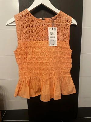 Zara Persikofärgad spetsblus med volang storlek S - Supersöt persikofärgad blus från Zara med spets upptill och smockad midja. Ärmlös modell med volang nedtill och rund halsringning. Perfekt för dig som gillar romantiska detaljer och en feminin look.