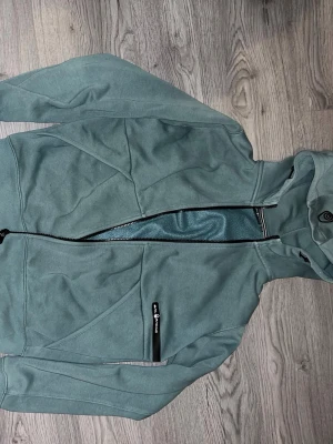 Grön hoodie från Peak Performance - Snygg grön hoodie från sail racing med dragkedja framtill och en praktisk bröstficka med svart dragkedja. Hoodien har huva med dragsko och är tillverkad i mjukt material, storlek 170 som motsvarar S. Säljer pga att den är för liten för mig, hör av dig vid frågor eller köp. Mvh Edvin
