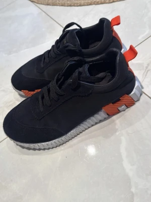 Svarta sneakers från h  med orange detaljer - Snygga svarta sneakers från h  med unika orange detaljer på hälen och sidorna. Skorna har en chunky vit sula med räfflat mönster och svarta skosnören. Ovandelen är i textil och mockaliknande material. Perfekta för dig som gillar streetstyle och vill sticka ut.