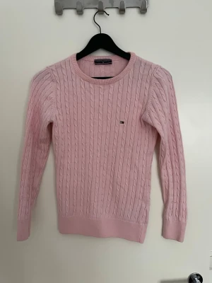 Ljusrosa kabelstickad tröja Tommy Hilfiger - Säljer en ljusrosa kabelstickad tröja från Tommy Hilfiger med rund halsringning och diskret logga på bröstet. Tröjan har långa ärmar och ribbade muddar vid ärmslut och nederkant. Perfekt för dig som gillar klassisk och stilren look.