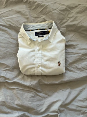 Vit skjorta från Polo Ralph Lauren - Klassisk vit skjorta från Polo Ralph Lauren med broderad logga på bröstet. Skjortan har lång ärm, knappar framtill och en snygg krage. Tillverkad i mjuk bomull för en clean och tidlös look. Skjortan är sparsamt använd därav i bra skick.