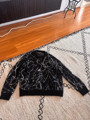 Svart stickad tröja med vitt mönster - Cool svart stickad tröja med abstrakt vitt mönster över hela plagget. Tröjan har rund halsringning och långa ärmar. Materialet ser mjukt och fluffigt ut, perfekt för dig som gillar en unik och konstnärlig stil.