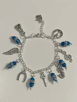 Silverfärgat berlockarmband med blå pärlor - HANDGJORT armband i silver med blå pärlor och flera olika berlocker. Armbandet har justerbar kedja.