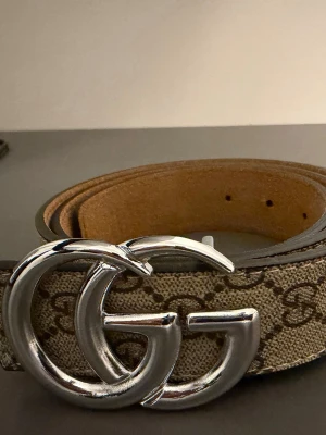 Gucci GG-bälte  - Snyggt beige bälte från Gucci med klassiskt GG-mönster i brunt och stor silverfärgad GG-spänne framtill. Bältet är gjort i canvas med läderdetaljer på insidan. Perfekt accessoar för att lyfta vilken outfit som helst.