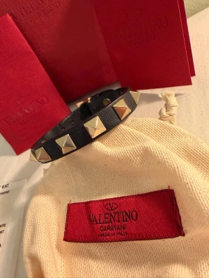 Valentino armband  - Snyggt svart armband från Valentino Garavani i äkta läder med ikoniska pyramidformade guldfärgade nitar. Justerbart spänne och exklusiv känsla. Kommer med originalpåse och ask, perfekt för dig som gillar edgy accessoarer. Med box och extra spikar. Lädret har gått upp lite men fixas lätt med lim