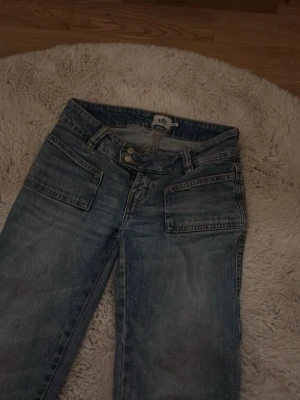 Blå bootcut jeans med stora fickor - Säljer ett par blå bootcut jeans med stora fyrkantiga framfickor och dubbla knappar i midjan. Jeansen har en tvättad look och raka ben med lätt utsvängning. 
