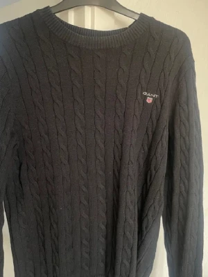 Svart kabelstickad tröja från GANT - Kabelstickad svart tröja från GANT med rund halsringning och diskret logga broderad på bröstet. Klassisk design med ribbade muddar vid hals, ärmslut och nederkant. Perfekt för dig som gillar stilrena och tidlösa plagg.